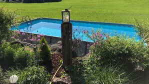 Een rechthoekig zwembad met een houten paal en lantaarn, omgeven door weelderige begroeiing en uitzicht op een landelijk landschap met huizen en heuvels.
