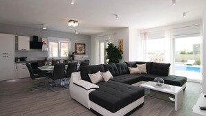 A modern living room with a sectional sofa, a dining area with a table and chairs, a kitchen with white cabinets, and a large window overlooking a swimming pool. - Beautiful home in Sukosan (Sukosan)