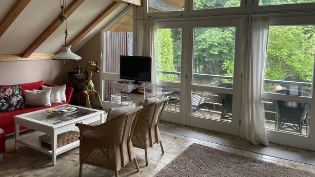 Ein geräumiges Wohnzimmer mit hoher Decke, großen Fenstern, einem Fernseher und einer gemütlichen Sitzecke.