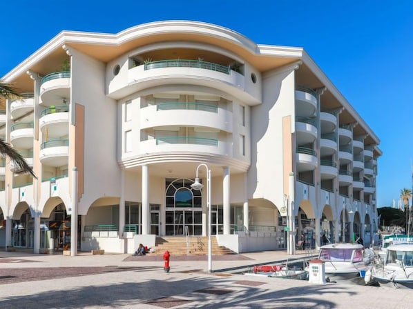 A modern hotel with multiple balconies, a central entrance with a glass facade, and a parking area with boats.