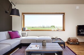 Ein modernes Wohnzimmer mit einer Eckcouch, einem Couchtisch, einem Billardtisch und einem großen Fenster mit Blick ins Freie.