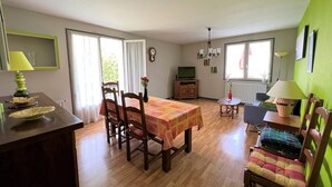 Ein Essbereich mit Tisch für zwei Personen, Holzsesseln, einem kleinen Beistelltisch mit Lampe und einem Fernseher im Hintergrund.