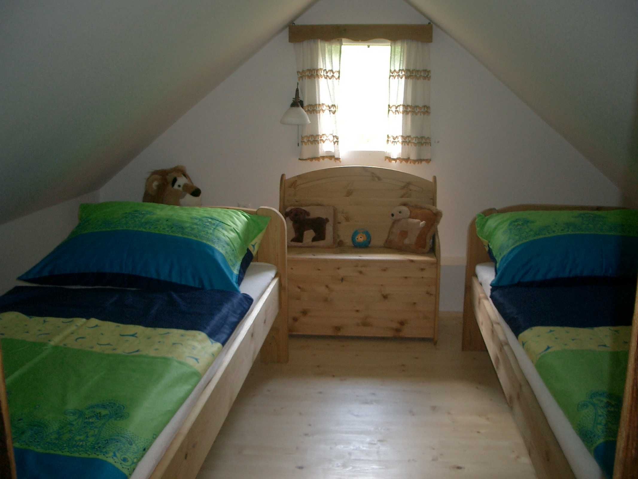 Une petite chambre mansardée avec deux lits simples, une commode en bois et une fenêtre avec des rideaux.