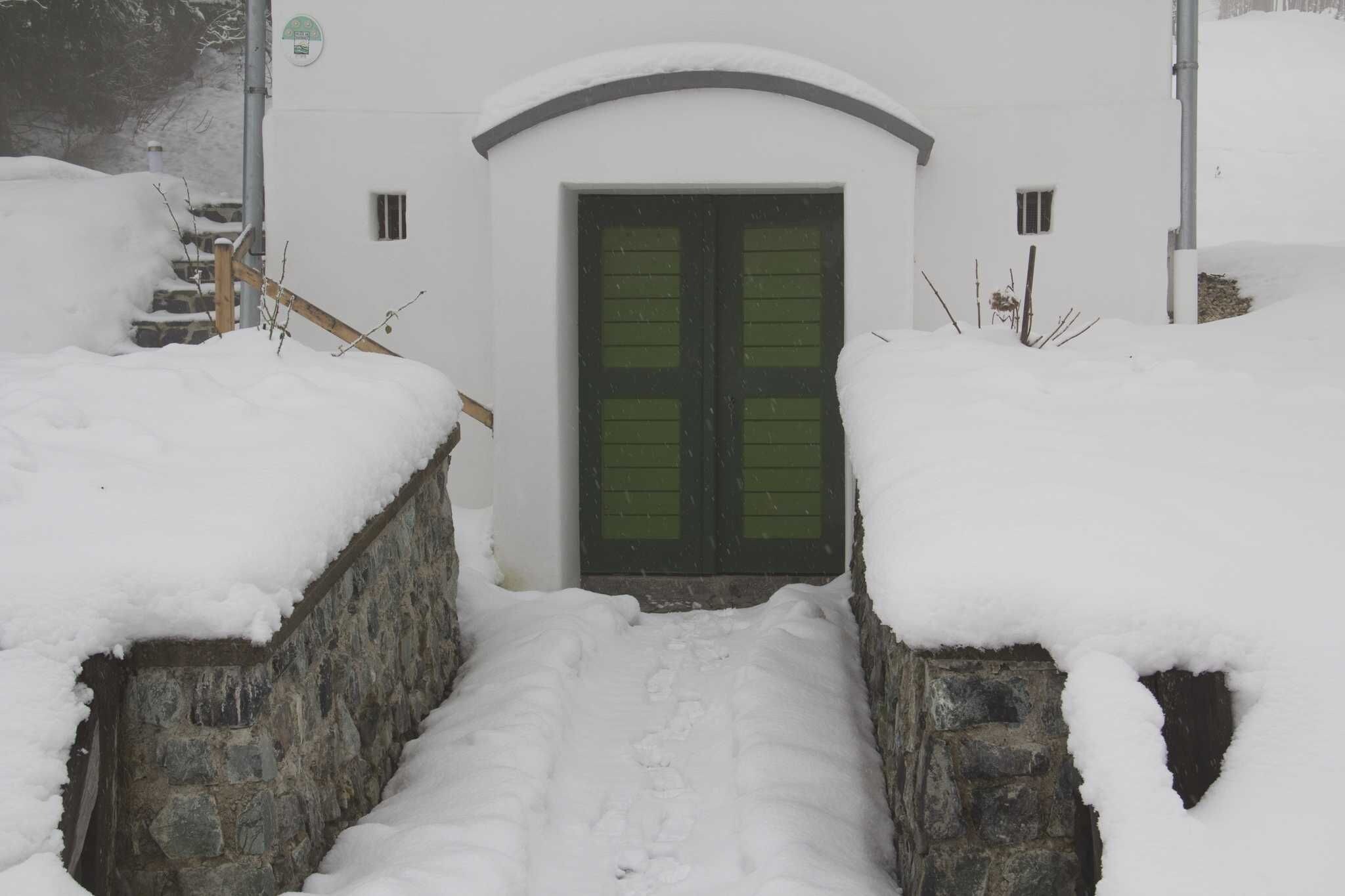 A snow-covered entrance with a stone wall, a wooden staircase, and a green door.