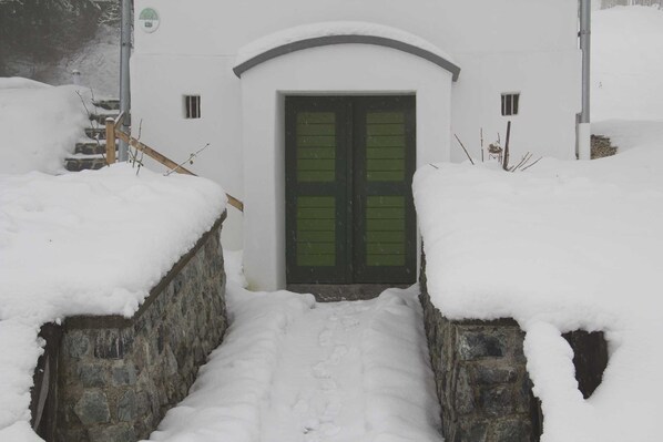 A snow-covered entrance with a stone wall, a wooden staircase, and a green door.
