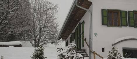 A snow-covered building with a number '1952' on the facade, a staircase leading to the entrance, and snow-laden trees and shrubs in the foreground.