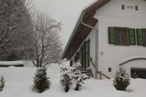 Un edificio cubierto de nieve con el número '1952' en la fachada, una escalera que conduce a la entrada, y árboles y arbustos cargados de nieve en primer plano.