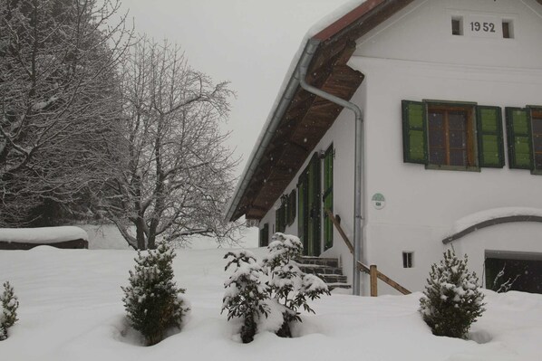 A snow-covered building with a number '1952' on the facade, a staircase leading to the entrance, and snow-laden trees and shrubs in the foreground.