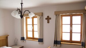 Un dormitorio con dos ventanas, cabecera de madera, mesita de noche y una cruz en la pared.