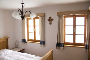 Un dormitorio con dos ventanas, cabecera de madera, mesita de noche y una cruz en la pared.