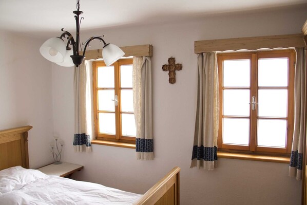 A bedroom with two windows, a wooden headboard, a bedside table, and a cross on the wall.