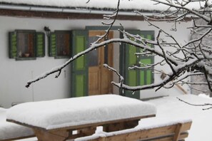 A snow-covered outdoor area with a wooden bench and a building with green shutters in the background.