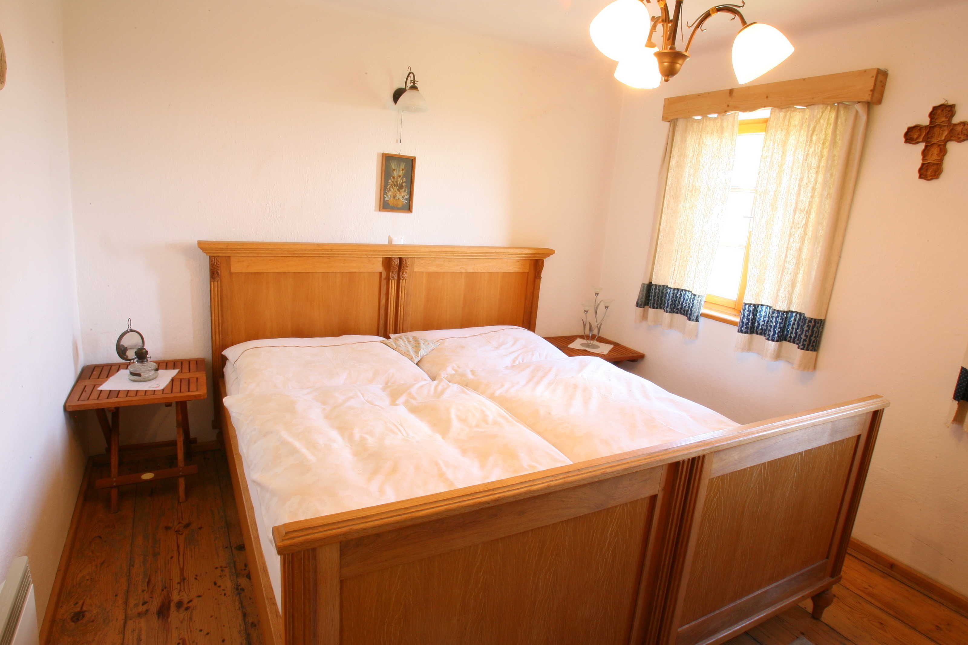 A bedroom with a wooden bed, a nightstand, a wall-mounted lamp, a window with curtains, and a cross on the wall.
