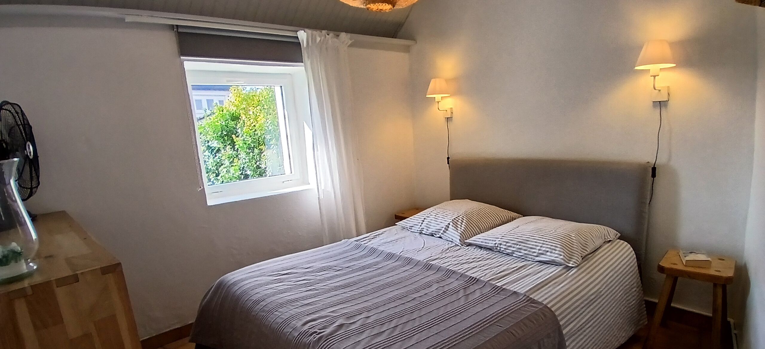A bedroom with a bed, two bedside tables, a lamp, a window with curtains, and a view of greenery outside.