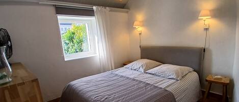 A bedroom with a bed, two bedside tables, a lamp, a window with curtains, and a view of greenery outside.