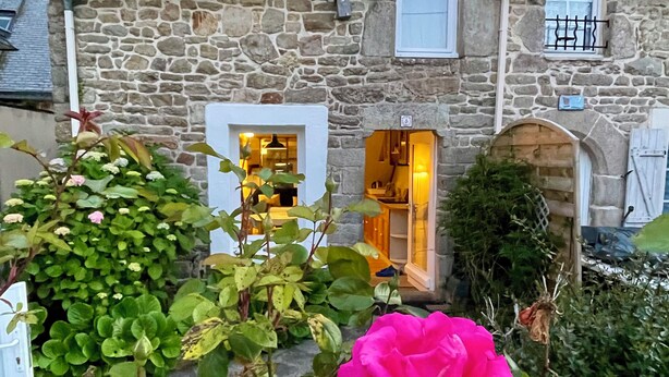 Un edificio de piedra con un jardín delantero, que incluye una rosa rosa y plantas verdes.