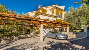 Un edificio de dos plantas con un coche estacionado frente a él, una pérgola cubierta de hiedra y un muro de piedra.