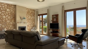 A living room with a stone wall, a fireplace, a sofa, a TV, and a view of the sea.