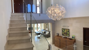 A modern interior with a staircase leading to a second floor, a large chandelier, a sofa, and a wooden cabinet with a painting.