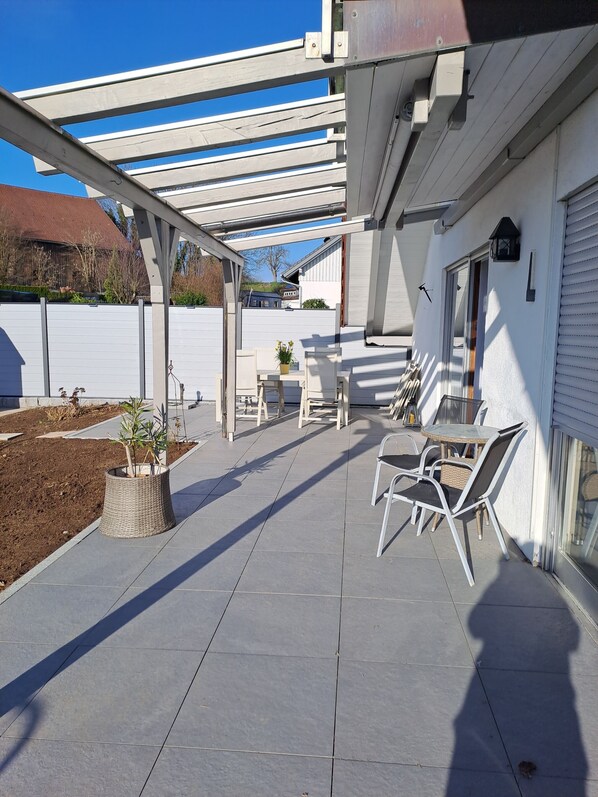 A covered patio area with a table and chairs, a potted plant, and a view of a building in the background.