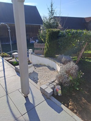 A paved patio area with a stone wall, a wooden bench, and a garden with various plants.