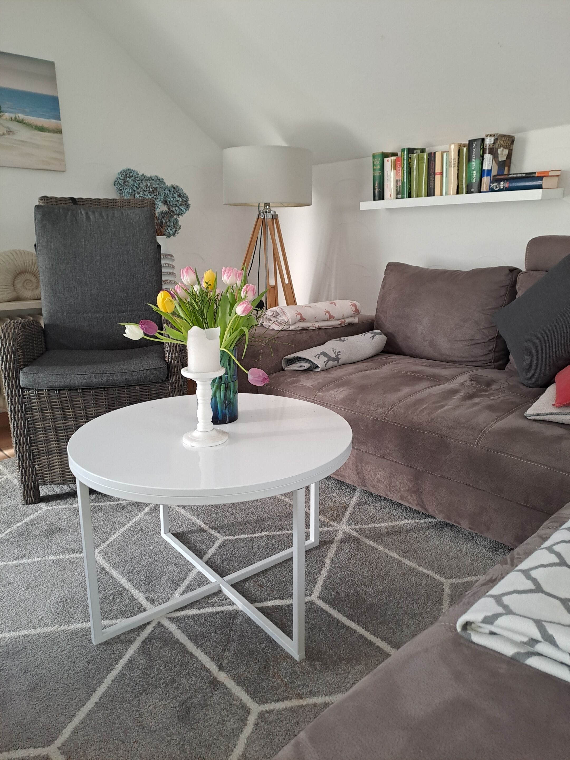 Ein Wohnzimmer mit einem runden weißen Tisch, einem braunen Sofa, einem Korbstuhl, einem Bücherregal und einem Gemälde an der Wand.