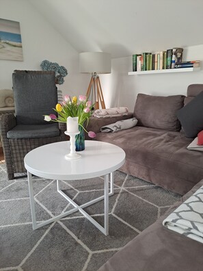 Una sala de estar con una mesa redonda blanca, un sofá marrón, un sillón de mimbre, una estantería y un cuadro en la pared.
