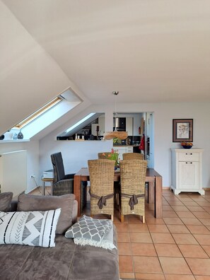A cozy living room with a brown sofa, a dining table with wicker chairs, a kitchen in the background, and a skylight above.