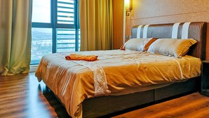 A hotel room with a large bed, a nightstand, a wall-mounted lamp, and a view of the city through large windows. - Green Haven by Tove Global (Masai)