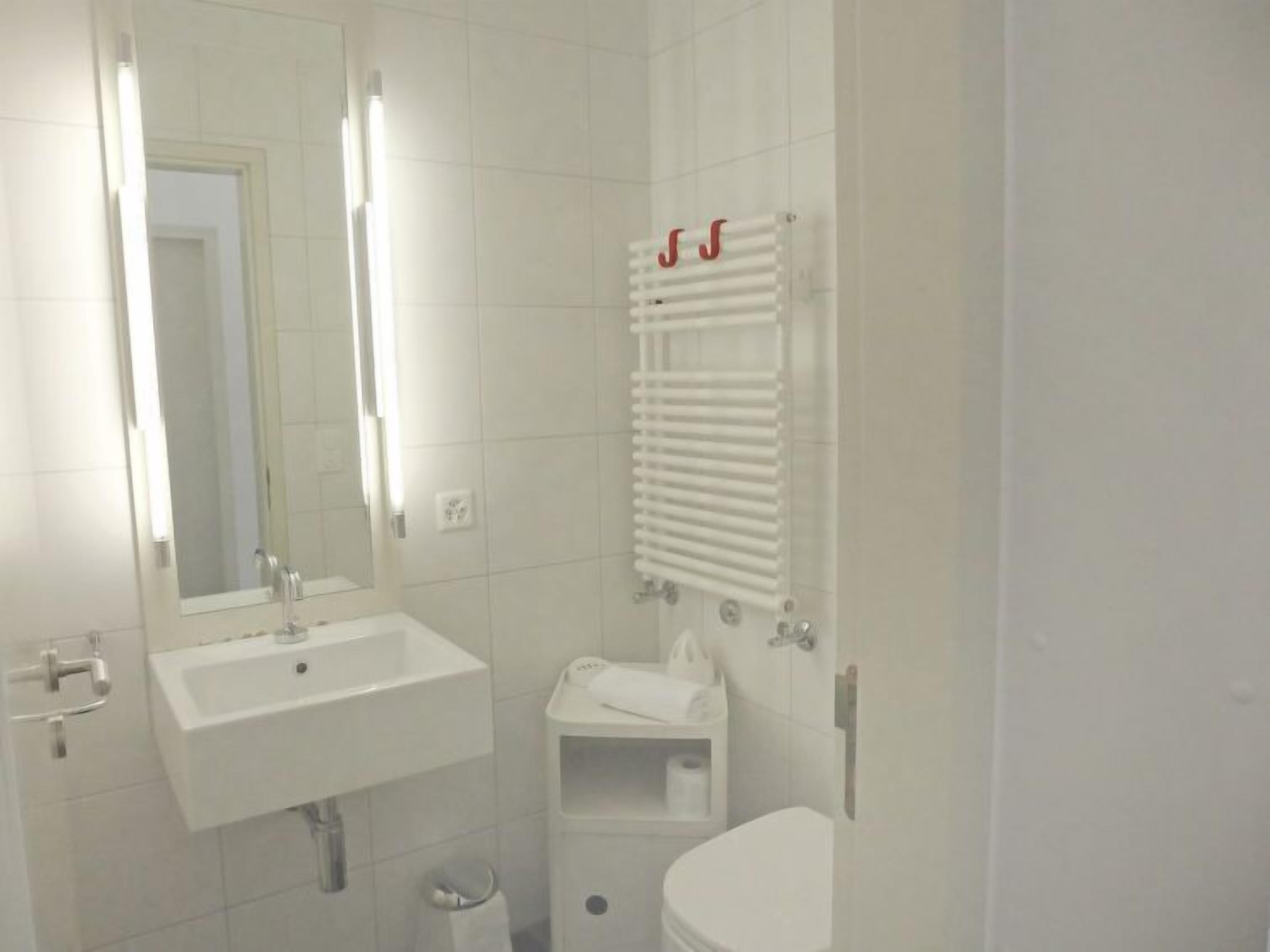 Ein Badezimmer mit weißem Waschbecken, Spiegel und Toilette. Ein Handtuchhalter mit Handtüchern und ein Mülleimer sind vorhanden.