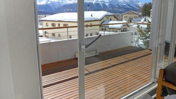 Een balkon met houten vloer en uitzicht op besneeuwde bergen en gebouwen.