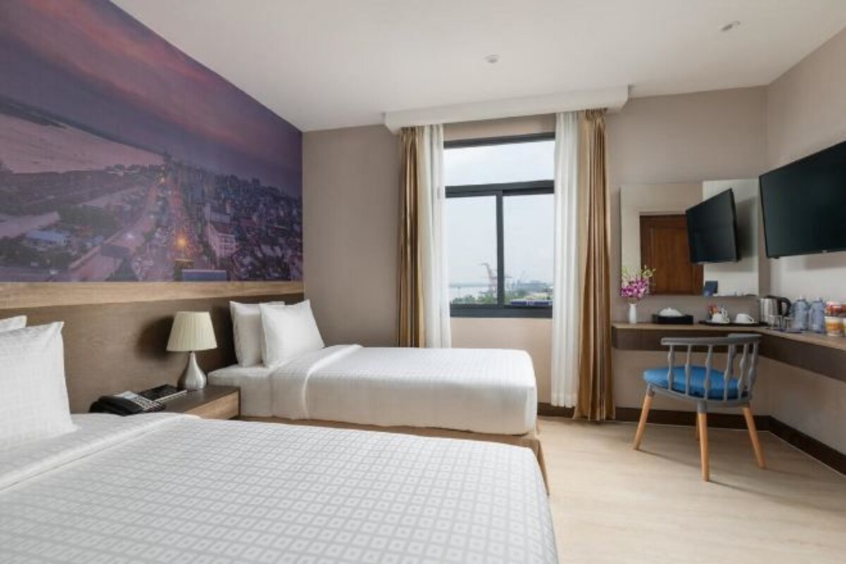 A hotel room with two beds, a desk, a chair, a TV, and a large window with a cityscape view.
