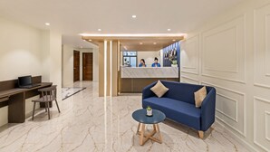 A modern lobby with a reception desk, a blue sofa, a small round table with a plant, and a marble floor. - Hotel Balmi (Yangon)