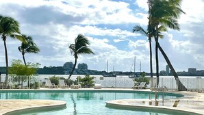 A swimming pool with a curved design, surrounded by palm trees and lounge chairs, with a marina and buildings in the background. - Lagoon front Magnificent Studio completely renovated with panoramic lagoon view (Saint-Martin)
