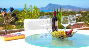 Una mesa con un bol de vidrio lleno de fruta, dos copas de vino, una botella de vino y un libro sobre una mesa de vidrio, con una vista de montañas al fondo.