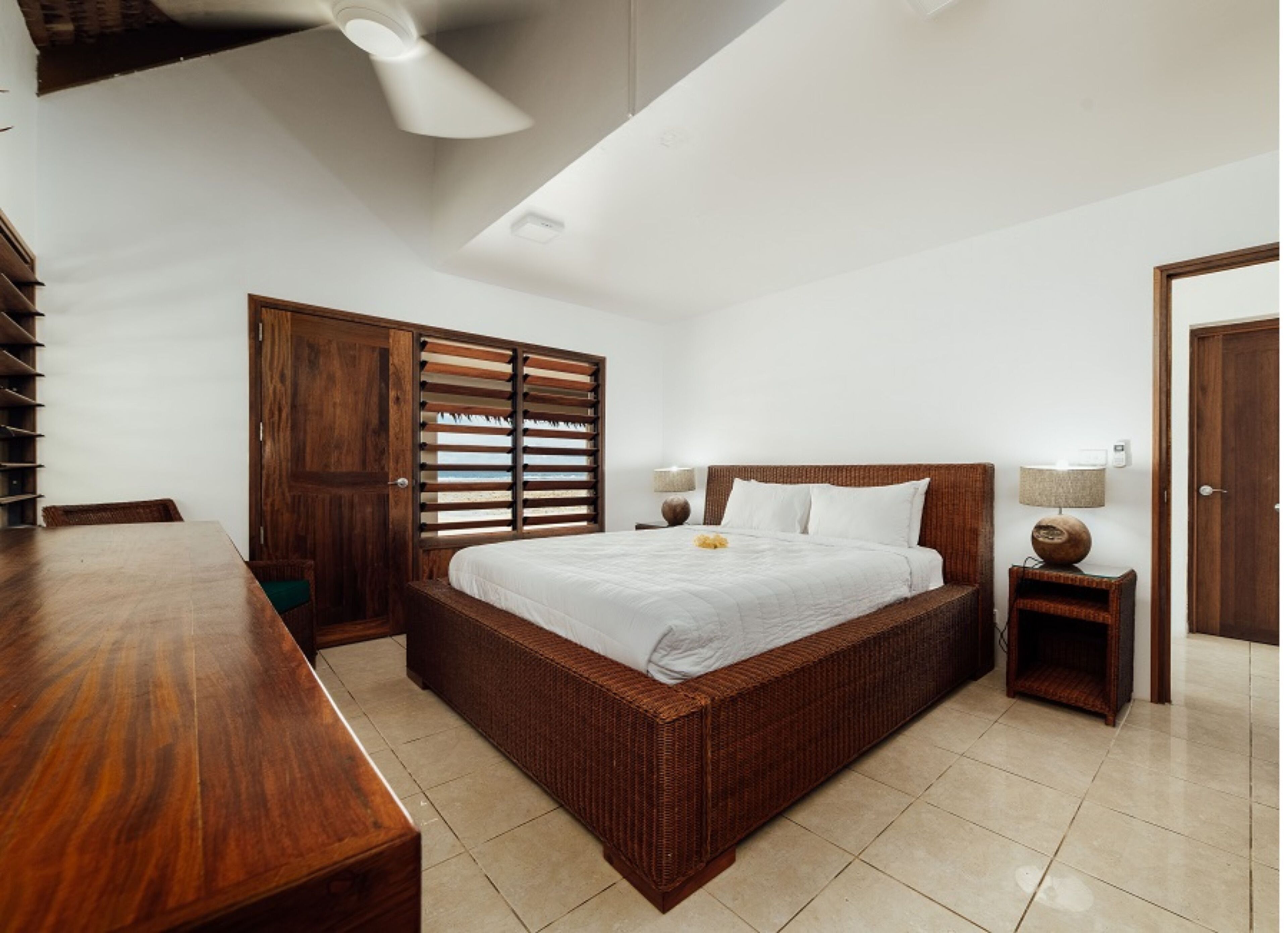A bedroom with a large bed, wooden furniture, a ceiling fan, and a view through the window blinds.