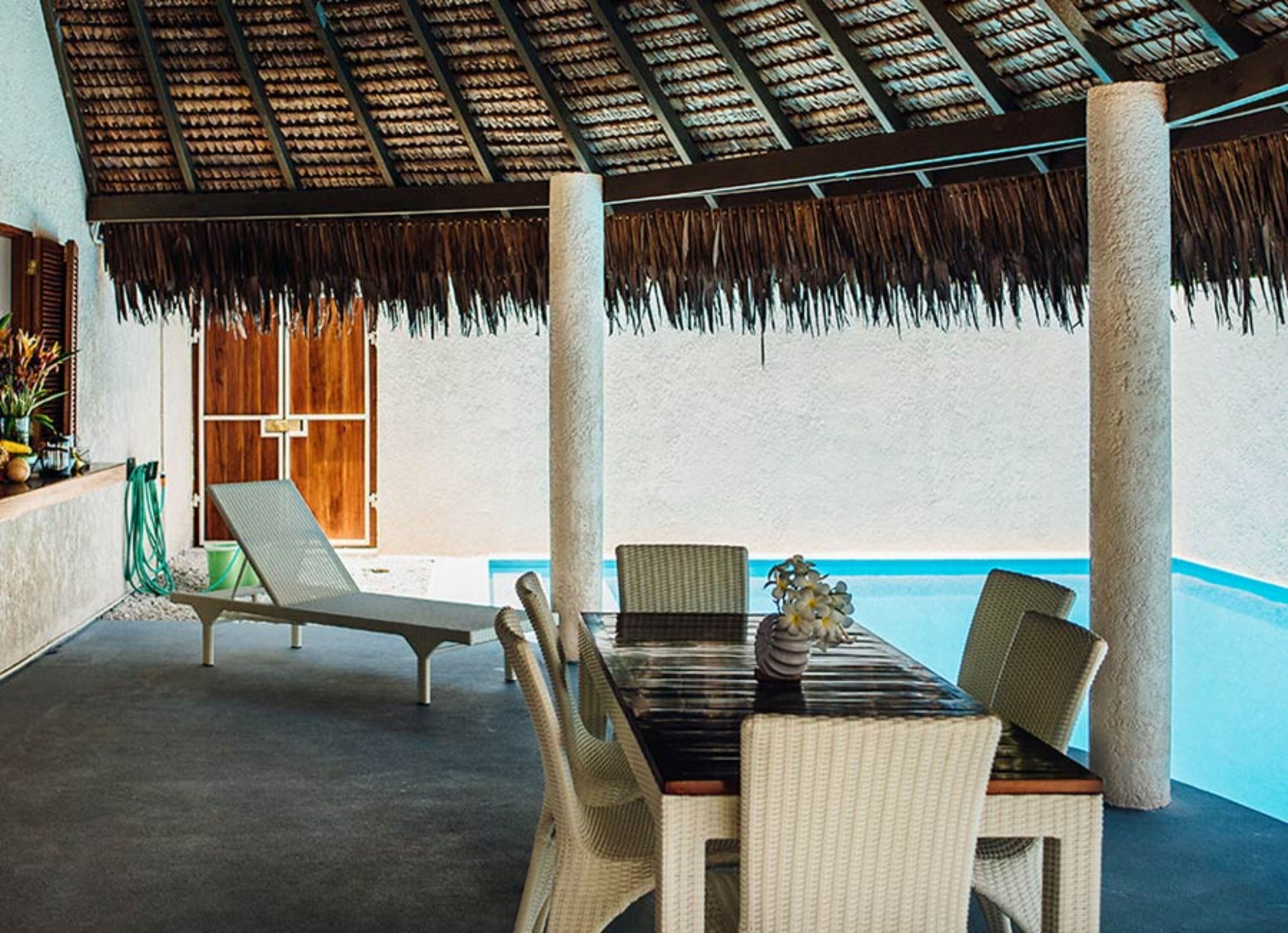 A covered outdoor seating area with a table and chairs, a pool in the background, and a thatched roof.