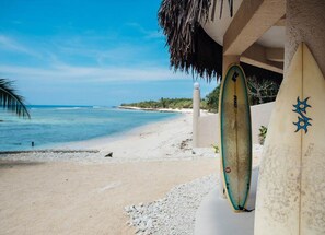 A beach view with a thatched roof structure, a surfboard leaning against it, and a clear blue sky. - Waves at Surfside (Port Vila)