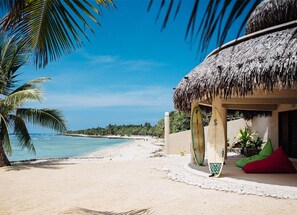A beach scene with a thatched-roof hut, a palm tree, and a surfboard leaning against the hut. - Waves at Surfside (Port Vila)