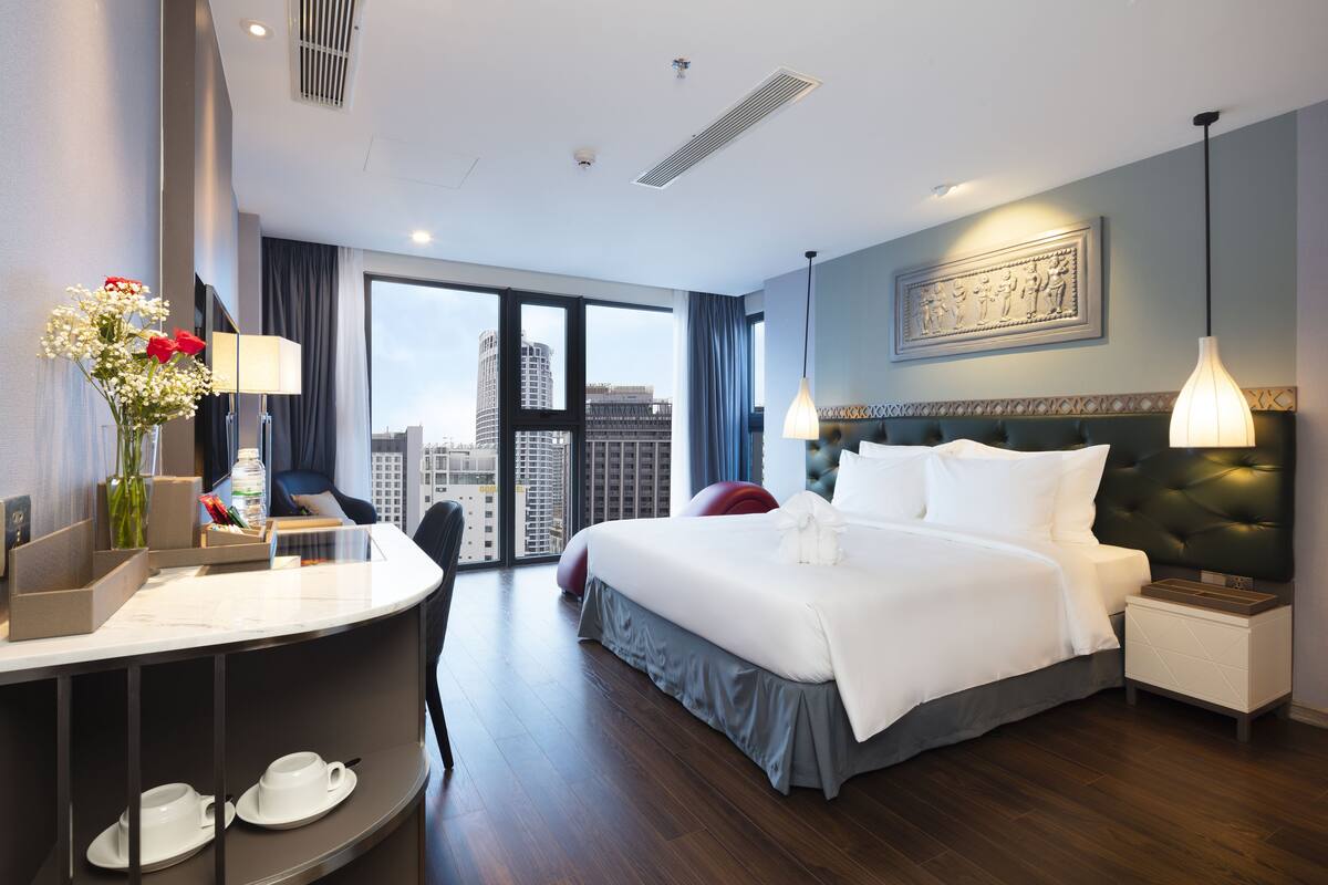 Ein Hotelzimmer mit einem großen Bett, einem Schreibtisch mit Stuhl, einem kleinen Tisch mit Tassen und Blick auf die Stadt durch große Fenster.