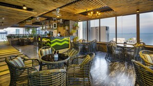A rooftop bar with wicker furniture, a bar counter, and large windows overlooking the city. - Erica Nha Trang Hotel (Nha Trang)
