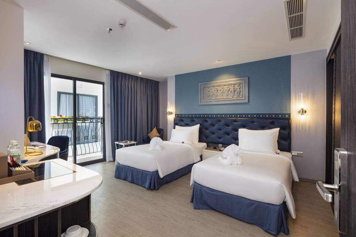 Ein Hotelzimmer mit zwei Betten, einem Schreibtisch, einem Sessel und einem Balkon mit Blick sowie einem Wandbild.