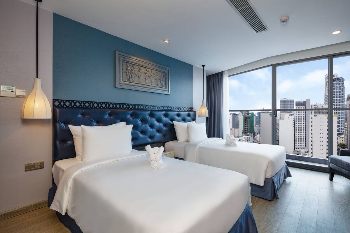 Ein Hotelzimmer mit zwei Betten, einem großen Fenster mit Blick auf die Stadt, einem dekorativen Wandbild und einer modernen Leuchte.