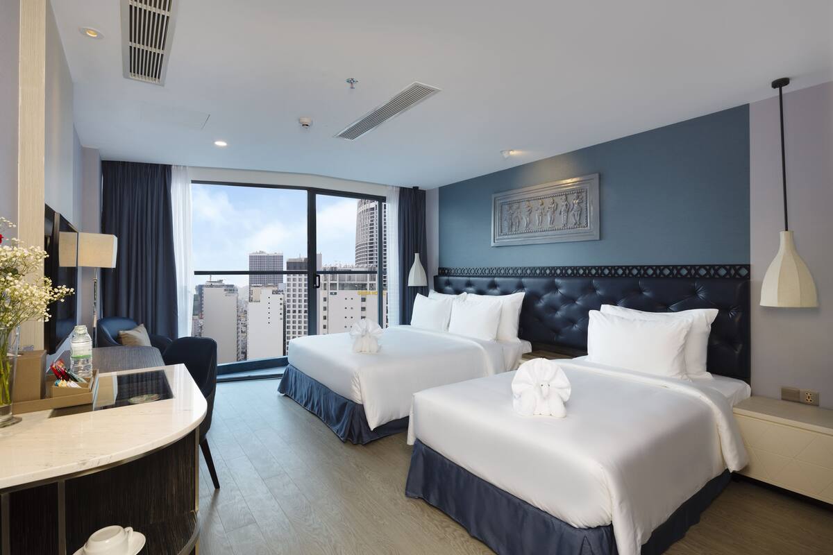 Ein Hotelzimmer mit zwei Betten, einem Schreibtisch, einem Stuhl, einem großen Fenster mit Blick auf die Stadt und einem gerahmten Kunstwerk an der Wand.