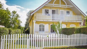 Une maison jaune à deux étages, avec une clôture blanche en piquets, un balcon et un petit jardin.
