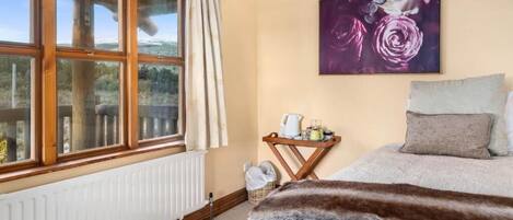 A bedroom with a large window, a painting of roses, a bed with a fur throw, and a small table with a kettle.
