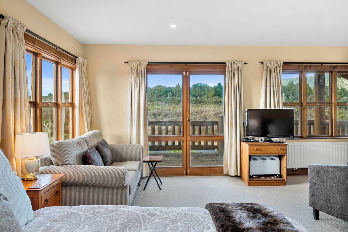 Een ruime kamer met een groot bed, een bank, een televisie op een houten dressoir en uitzicht op het landschap door grote ramen.