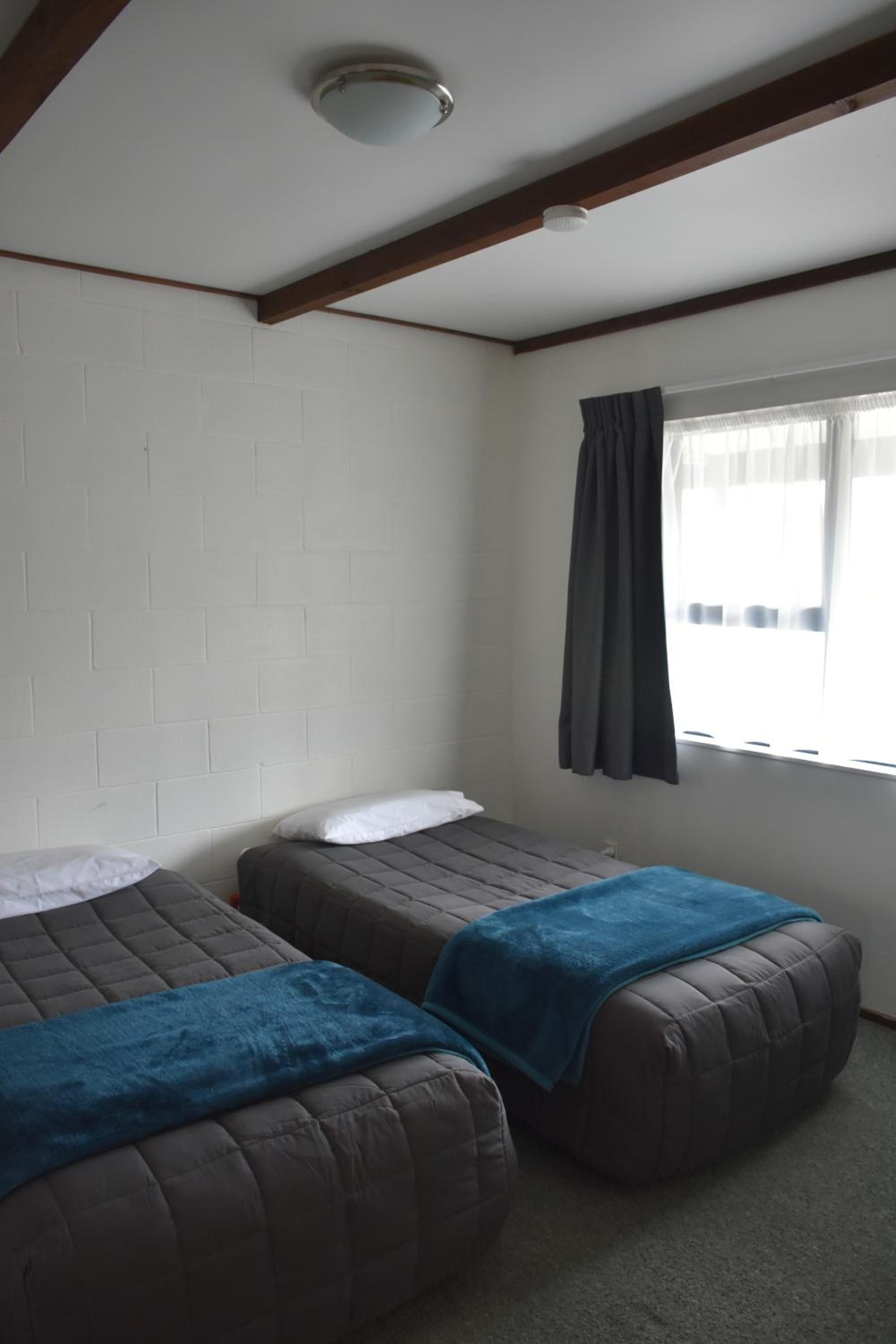 A room with two beds, a window with curtains, a white brick wall, and a ceiling light.