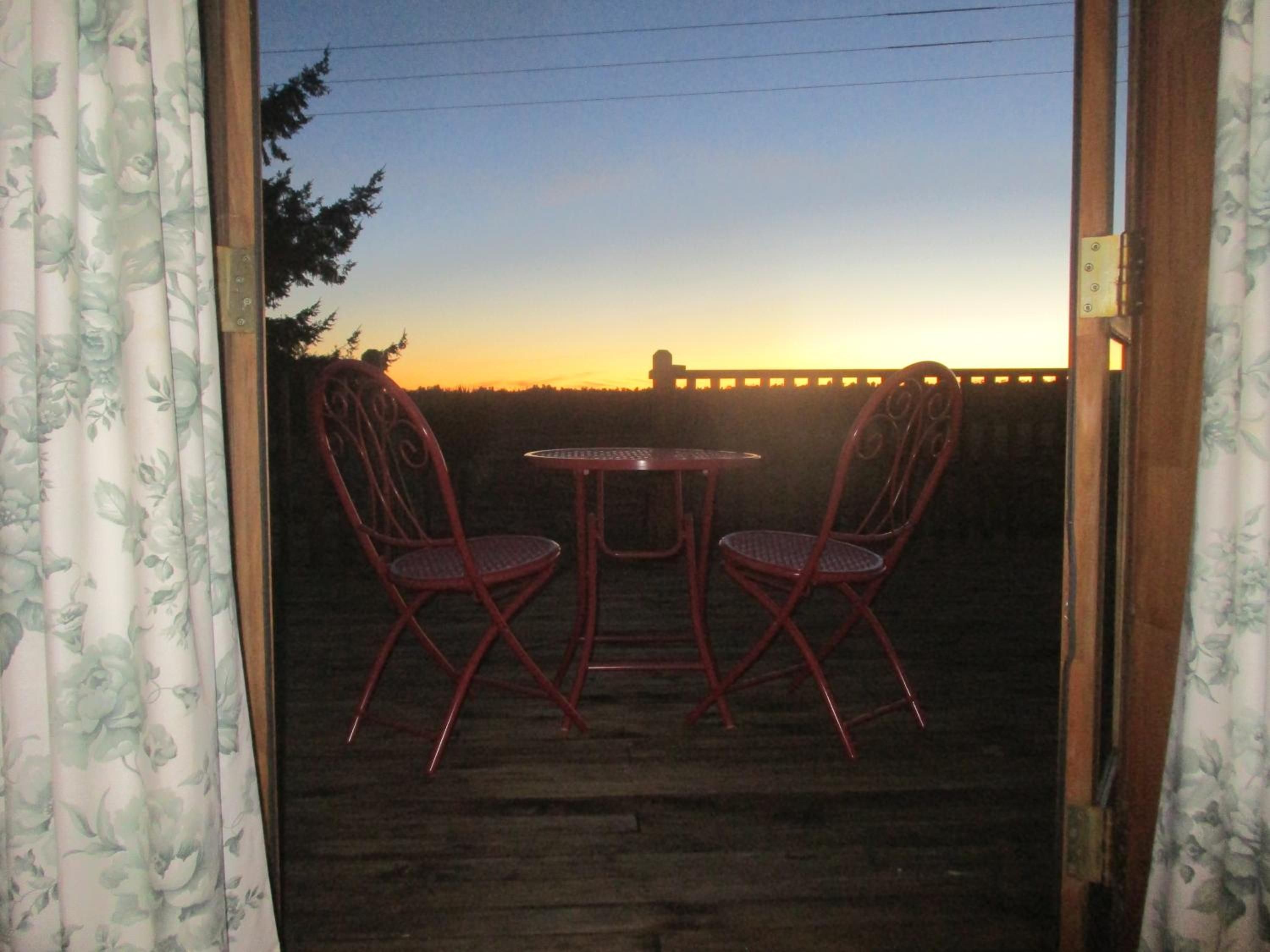 An outdoor patio with a table and two chairs, a wooden deck, and a view of the sunset.
