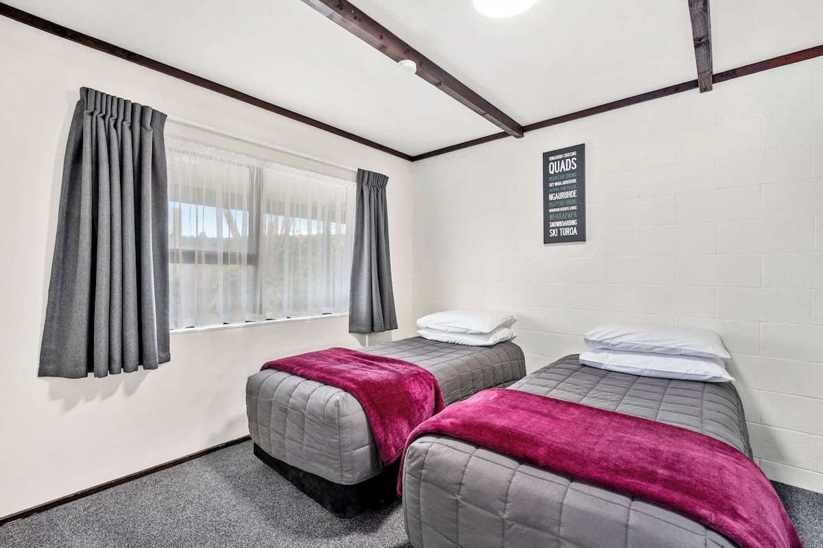 Een kamer met twee bedden, grijs beddengoed, een rode deken, een raam met grijze gordijnen en een bord met een overzicht van de kamertypes.